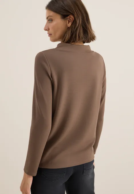 Frau mit braunem Pullover und dunkler Hose blickt nach links, Rückenansicht.
